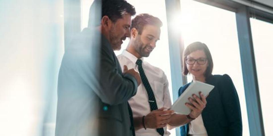 Gruppe von Geschäftsleuten, die gemeinsam ein digitales Tablet vor den Fenstern eines Bürogebäudes mit Blick auf die Stadt benutzen